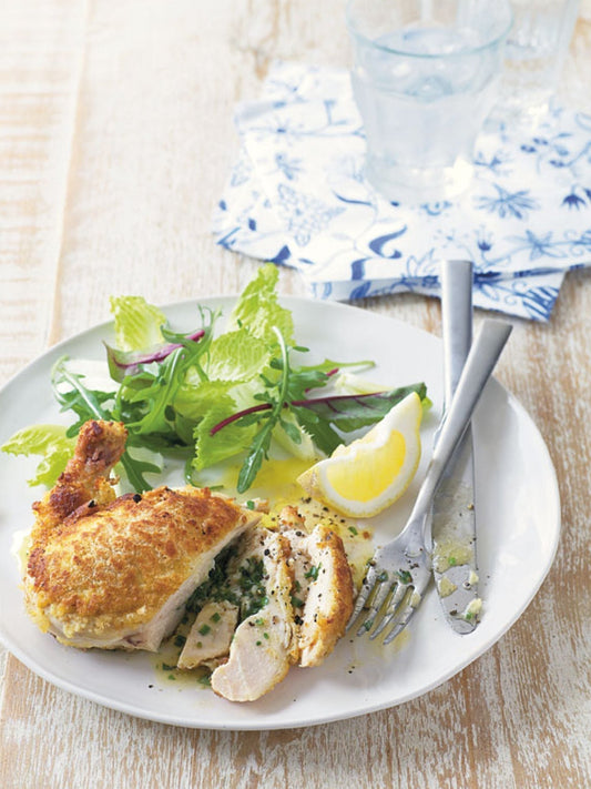 Knuspriges Chicken Kiev auf einem weißen Teller mit frischem Blattsalat und Zitronenspalte, serviert auf hellem Holztisch – ein saftiges Hähnchengericht mit Kräuterbutter-Füllung und goldbrauner Panade.