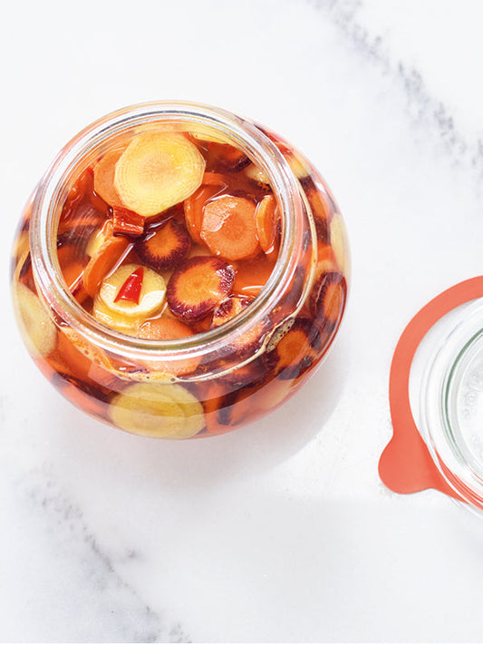 t: Geöffnete Einmachglas mit bunten, in Salzlake fermentierten Möhrenscheiben und Chilistücken auf hellem Untergrund, daneben liegender Gummiring-Deckel.