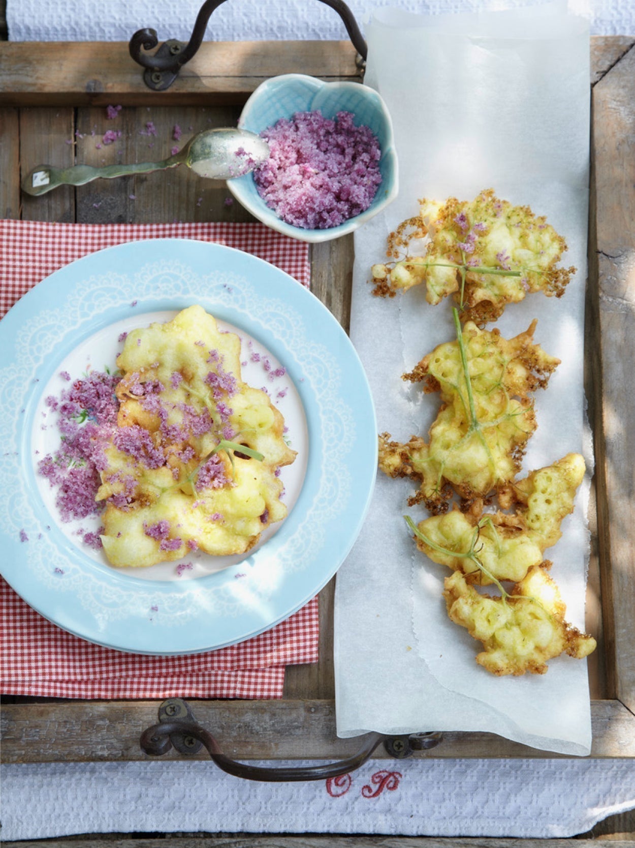 Gebackene Holunderblüten auf Servierbrett mit blauen Tellern, bestreut mit Veilchenzucker, daneben Schälchen mit violettem Blütenzucker.