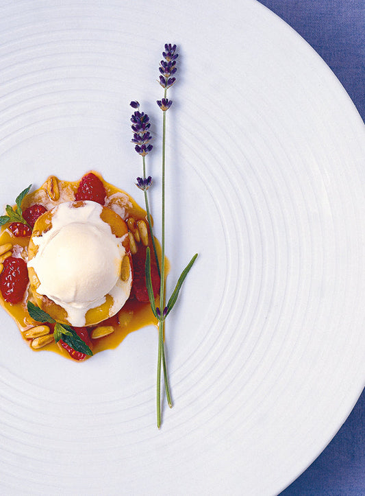 Gebratener weißer Pfirsich mit einer Kugel Lavendeleis, umgeben von Himbeeren, Pinienkernen und Sirup, dekoriert mit frischem Lavendel auf einem weißen Teller.