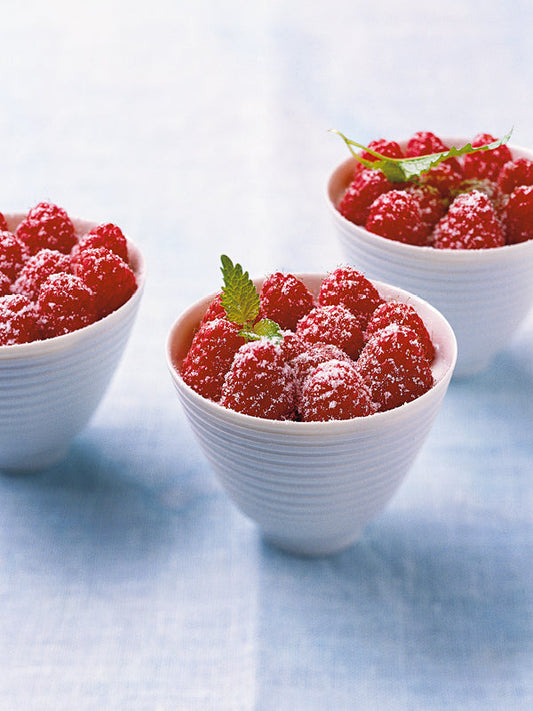 Drei weiße Schalen mit frischen Himbeeren, bestäubt mit Puderzucker und dekoriert mit Zitronenmelisse, angerichtet auf hellblauer Tischdecke.