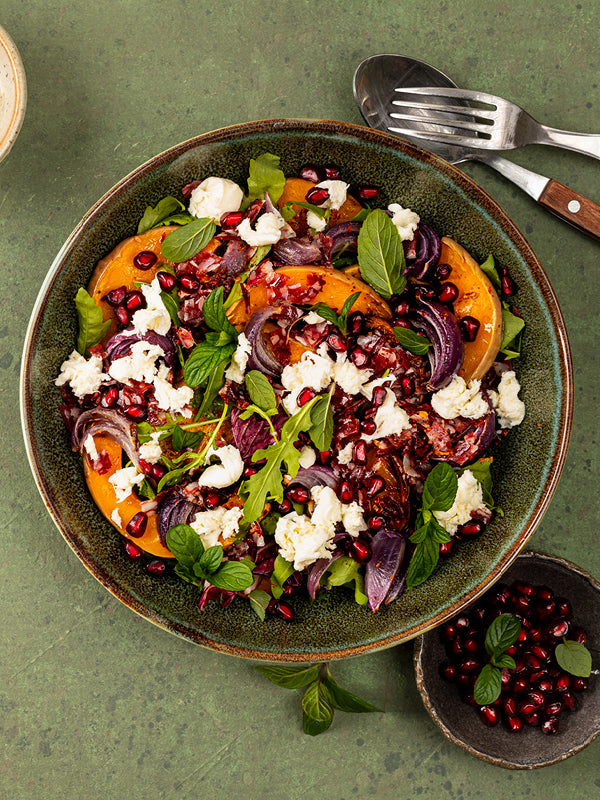 Herbstlicher Salat mit geröstetem Kürbis, roten Zwiebeln, Rucola, zerkrümeltem Feta, Granatapfelkernen und frischen Kräutern in einer grünen Keramikschale auf grünem Hintergrund
