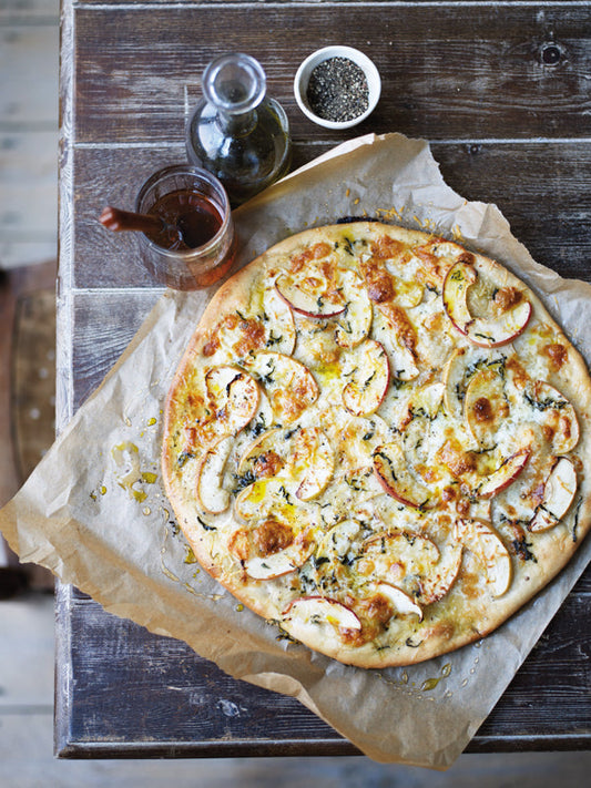 Knusprige Apfel-Taleggio-Tarte auf Backpapier, belegt mit Apfelscheiben, geschmolzenem Käse, Salbei und Olivenöl, auf einem rustikalen Holztisch.