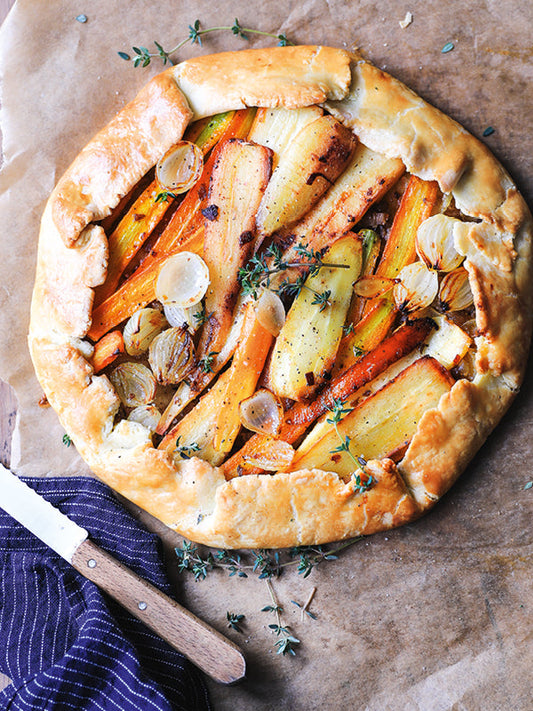 Rustikale Pastinaken-Karotten-Tarte auf Backpapier, belegt mit geröstetem Wurzelgemüse, Perlzwiebeln und Maronen, dekoriert mit frischem Thymian.