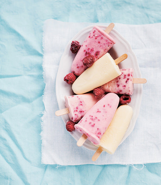 Hausgemachtes Himbeer-Vanille-Eis am Stiel in einer weißen Schale, garniert mit frischen Himbeeren auf einem hellblauen Tuch.
