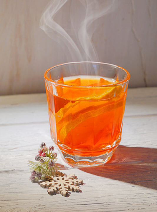 Ein dampfender Hot Aperol in einem Glas mit Orangenscheiben auf hellem Holz, daneben ein kleines weihnachtliches Deko-Element aus Beeren und Schneeflocke.