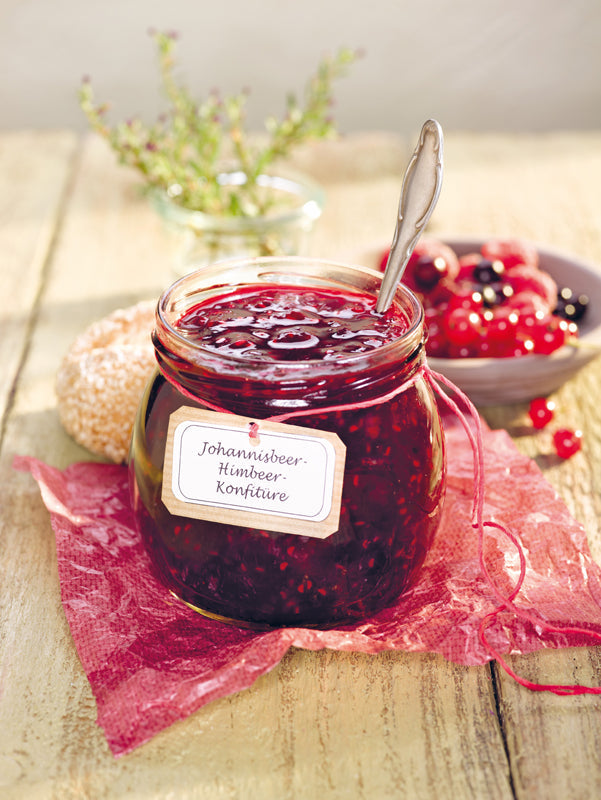 Offenes Glas mit selbstgemachter Johannisbeer-Himbeer-Konfitüre, dekoriert mit Schleife und Etikett, auf rosafarbenem Papier – im Hintergrund frische Beeren, Brötchen und Kräuter auf einem Holztisch.