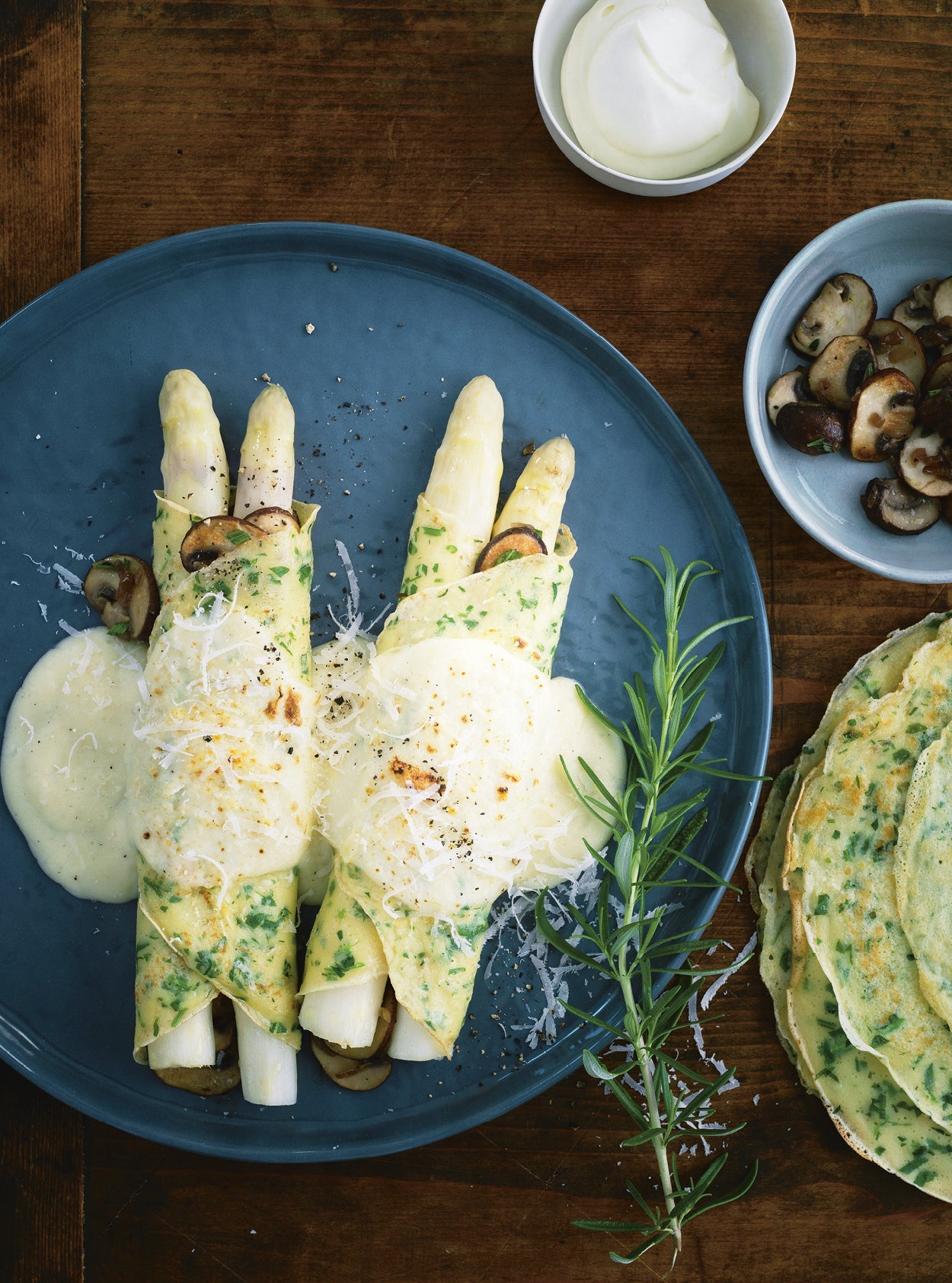 Gefüllte Kräuterpfannkuchen mit weißem Spargel, Pilzen und cremiger Béchamelsauce auf einem Teller, garniert mit Parmesan und frischem Rosmarin.