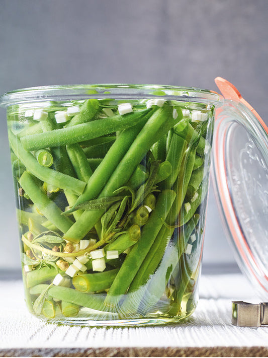 Nahaufnahme eines Einmachglases mit grünen Bohnen, frischem Bohnenkraut, klein gewürfeltem Meerrettich und eingelegten Pfefferkörnern in einem klaren Essigsud.