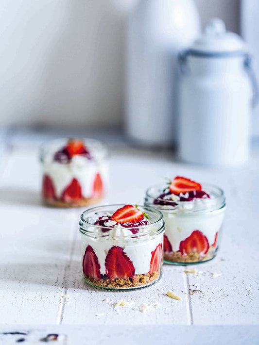 Dessertgläser mit New York Cheesecake im Glas, geschichtet mit Keksboden, frischen halbierten Erdbeeren, cremiger Frischkäse-Joghurt-Masse und fruchtigem Erdbeerpüree, garniert mit weißen Schokoraspeln und Erdbeerhälften.
