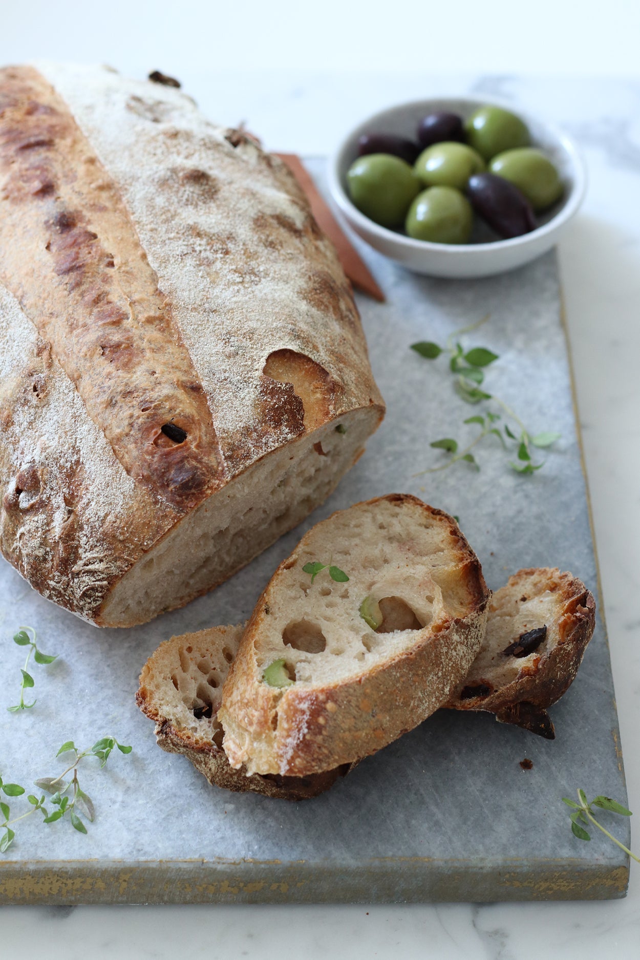 Rustikales Oliven-Parmesan-Brot mit knuspriger Kruste, angeschnitten auf einer Marmorplatte, dazu frischer Thymian und eine Schale mit grünen und schwarzen Oliven.