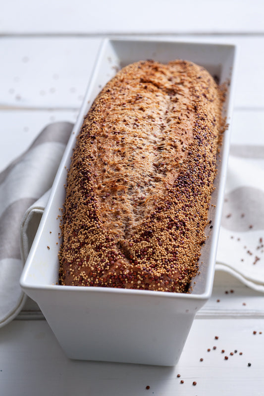 Frisch gebackenes Quinoa-Brot in einer weißen Kastenform, bestreut mit roten Quinoa- und Amaranthkörnern, auf einem weißen Holztisch mit gestreiftem Küchentuch.