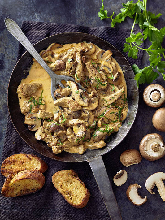 Eine Pfanne mit cremigem Rahmgeschnetzeltem und Champignons, bestreut mit frischer Petersilie; daneben geröstete Knoblauchbrotscheiben und frische Pilze.