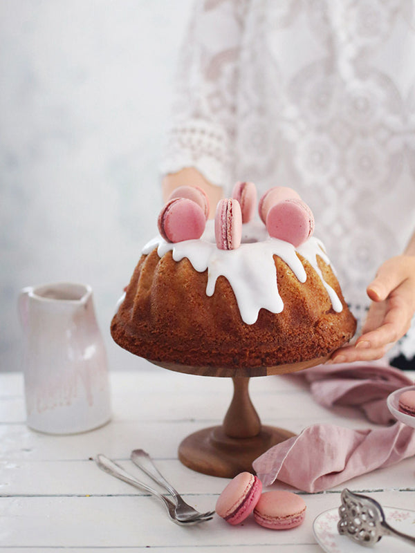 Saftiger Rhabarber-Marzipan-Gugelhupf mit weißem Zitronenguss und rosa Macarons, angerichtet auf einer Holz-Etagere vor heller Kulisse