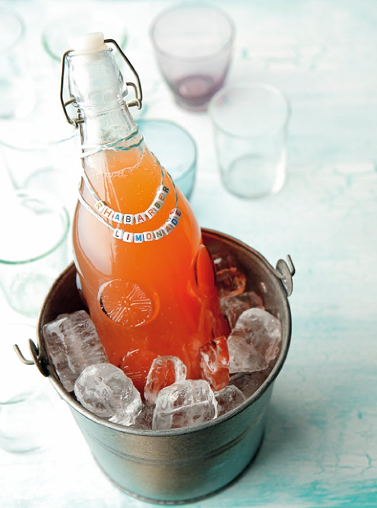 Hausgemachte Rhabarberlimonade in einer Glasflasche mit Bügelverschluss, gekühlt im Eiseimer, dekoriert mit Perlenkette und Schriftzug „Rhabarber Limonade“