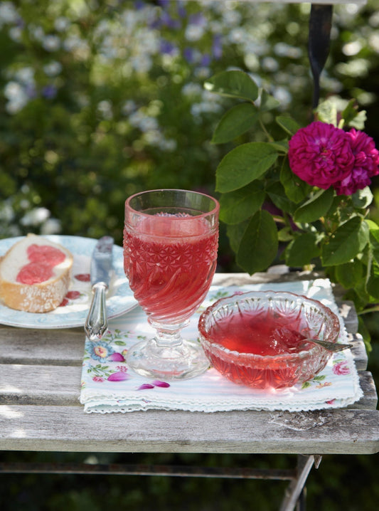 Rosenblüten-Gelee in Glas und Schale auf Gartentisch, mit Brot und blühender Duftrose im Hintergrund.