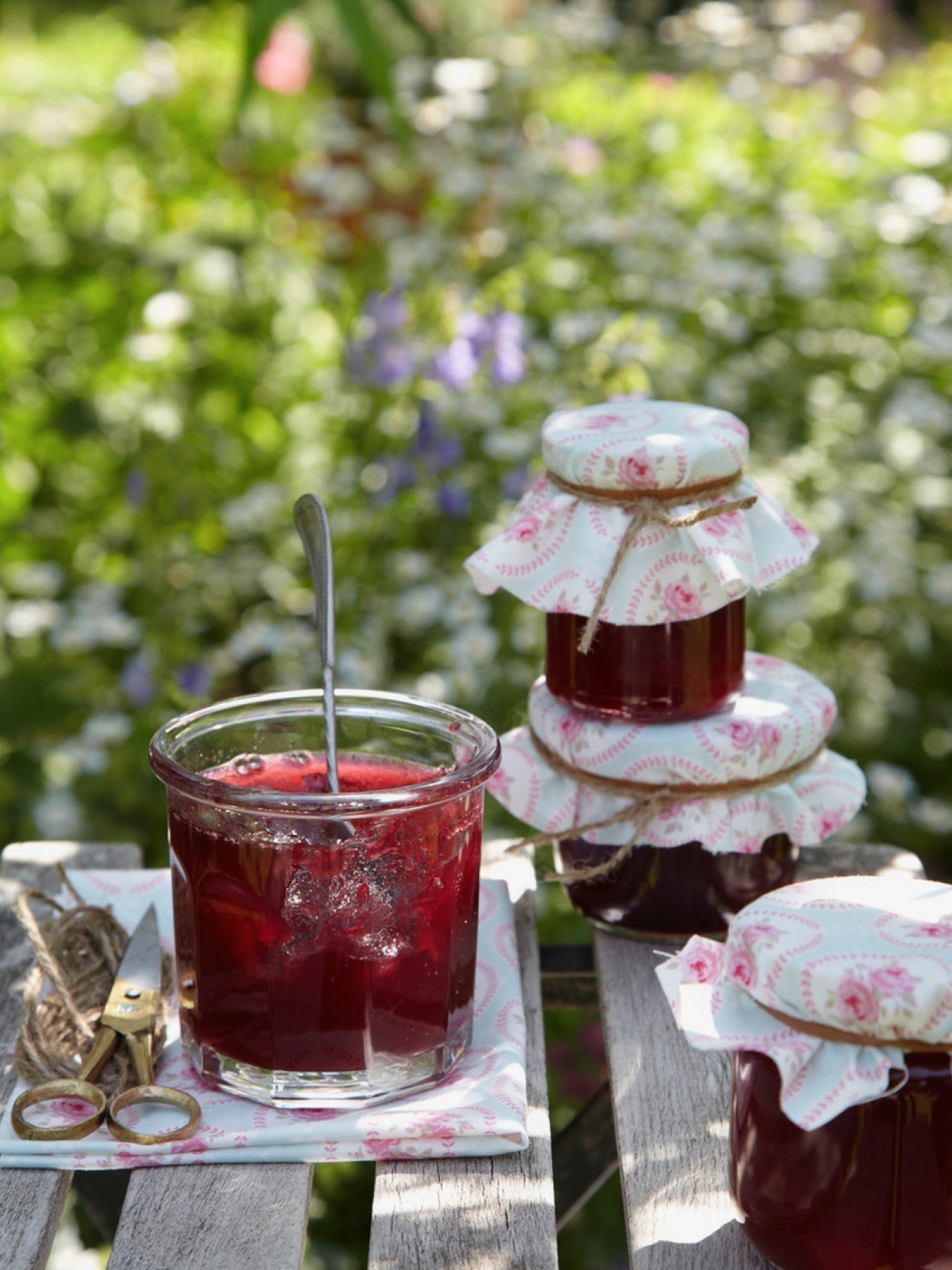 Tiefrote Sauerkirschmarmelade in Gläsern mit nostalgischem Stoffdeckchen, dekorativ auf Gartentisch mit Kordel und Schere arrangiert