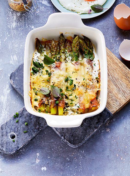 Überbackenes Spargel-Gratin mit grünem Spargel, Schinken, Käse und Kräutern in einer weißen Auflaufform auf rustikalem Untergrund – servierfertig und appetitlich angerichtet.