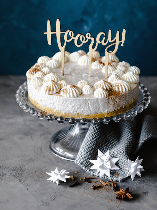 Ein Spekulatius-Cheesecake auf einer Glas-Tortenplatte, dekoriert mit Sahnetupfern und winterlichem Gewürz; davor Origami-Sterne und Anis auf grauem Untergrund.