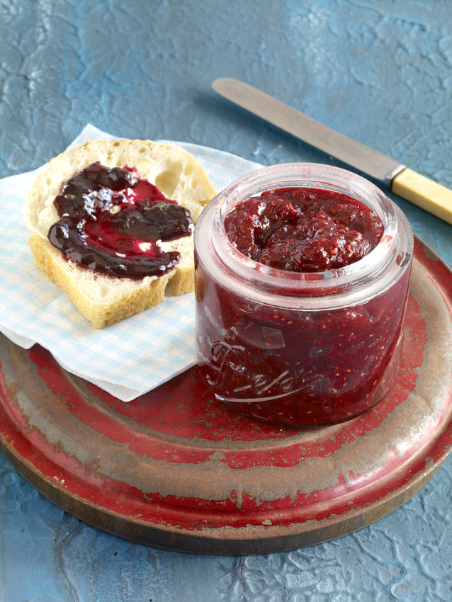 Ein geöffnetes Glas mit dunkler Stachelbeer-Himbeer-Konfitüre auf einem roten Teller, daneben eine Brotscheibe mit Butter und Konfitüre bestrichen – sommerlicher Fruchtaufstrich angerichtet auf blauem Hintergrund.