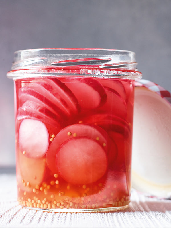 Nahaufnahme eines Glases mit süßsauer eingelegten Radieschenscheiben und Senfkörnern in klar-rotem Essigsud, dekorativ und appetitlich präsentiert auf hellem Untergrund