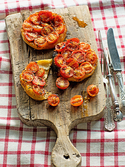 Herzhafte Mini-Tomaten-Tartes aus Blätterteig mit halbierten Kirschtomaten, Comté-Käse und Kräutern, angerichtet auf rustikalem Holzbrett mit Besteck – sommerliche Vorspeise auf rot-weiß karierter Tischdecke.