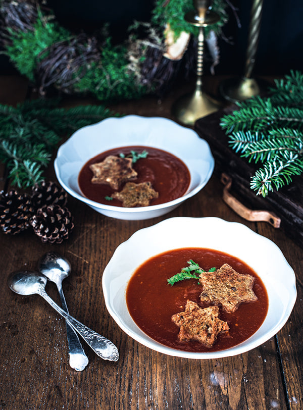 Zwei Schalen weihnachtliche Tomatensuppe, garniert mit knusprigen Sternen-Croutons, dekoriert mit Tannenzweigen und Zapfen auf einem Holztisch.