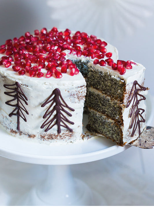 Eine mehrstöckige Wintertorte mit Mohnböden, weißer Creme, Schoko-Tannenbäumen und Granatapfelkernen obenauf; ein Stück ist herausgeschnitten und wird serviert.