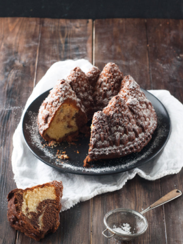 Ein Tannenbaum-Marmorkuchen auf einem dunklen Teller, leicht mit Puderzucker bestäubt, mit einem herausgeschnittenen Stück daneben; rustikale Holzoberfläche und feines Sieb im Vordergrund.