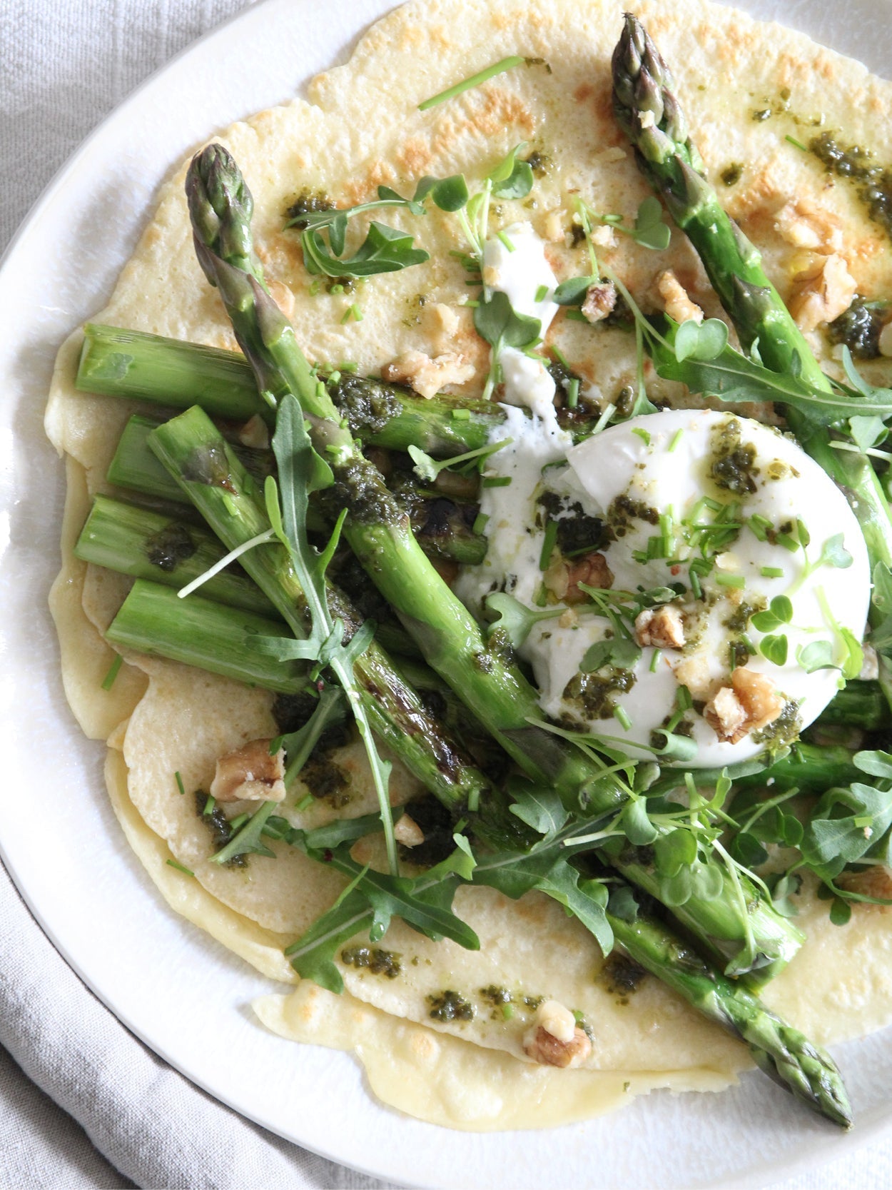 Herzhafter Pfannkuchen mit grünem Spargel, Burrata, Bärlauchpesto und frischen Kräutern, garniert mit Walnüssen auf einem hellen Teller.