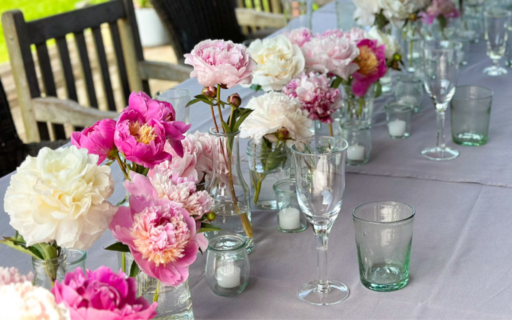 Lange festlich gedeckte Tafel im Freien mit einzelnen Pfingstrosen in kleinen Glasvasen, dekoriert in zarten Rosa-, Pink- und Cremetönen, ergänzt durch Kerzengläser und Trinkgläser.