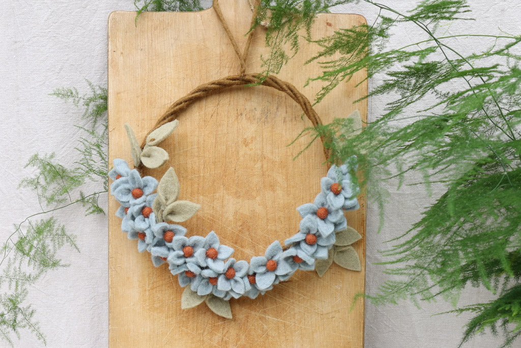 Filzkranz mit hellblauen Blüten an ein Brett gehangen.