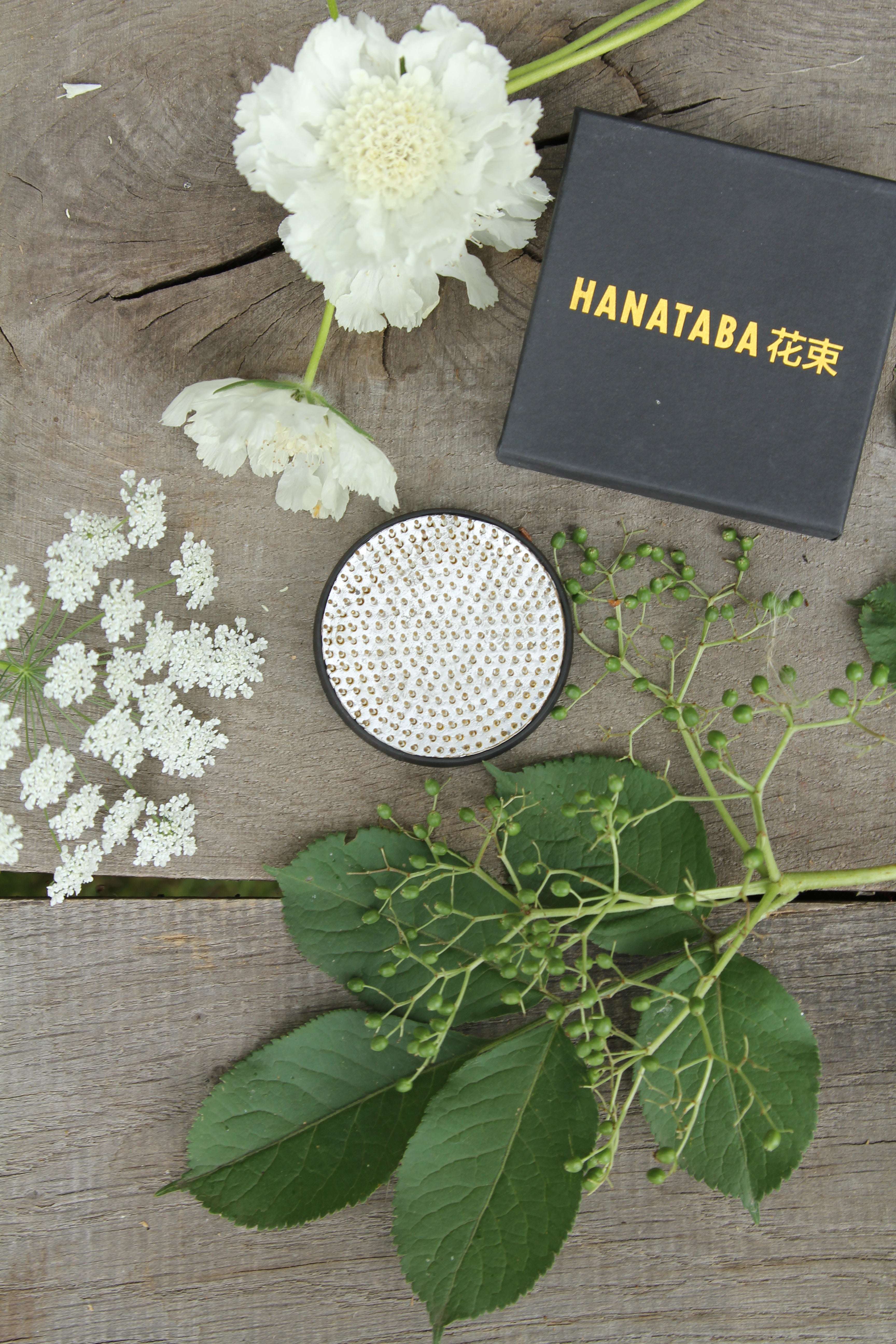 Elegante schwarze Box mit goldener Aufschrift "HANATABA", umgeben von grünen Blättern, zarten weißen Blumen auf rustikalem Holz.