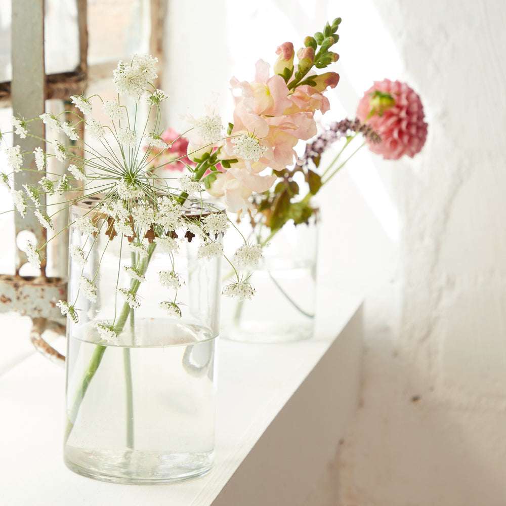 Zwei Glasvasen mit Blumenarrangements, darunter rosa, weiße und lila Blüten, auf Fensterbank neben altem, verrostetem Fensterrahmen.