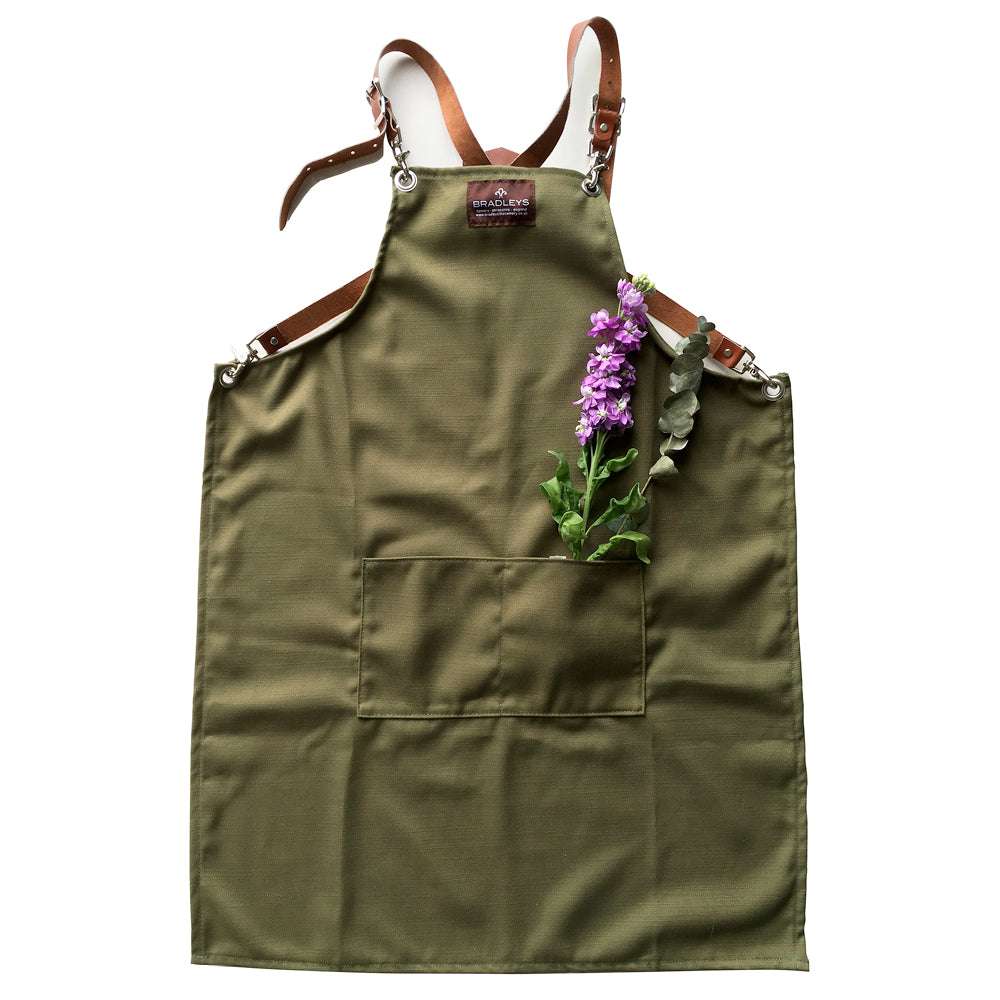 Grüne Küchenschürze mit braunen Lederriemen und Metallbeschlägen, violette Blume in der Vordertasche platziert.