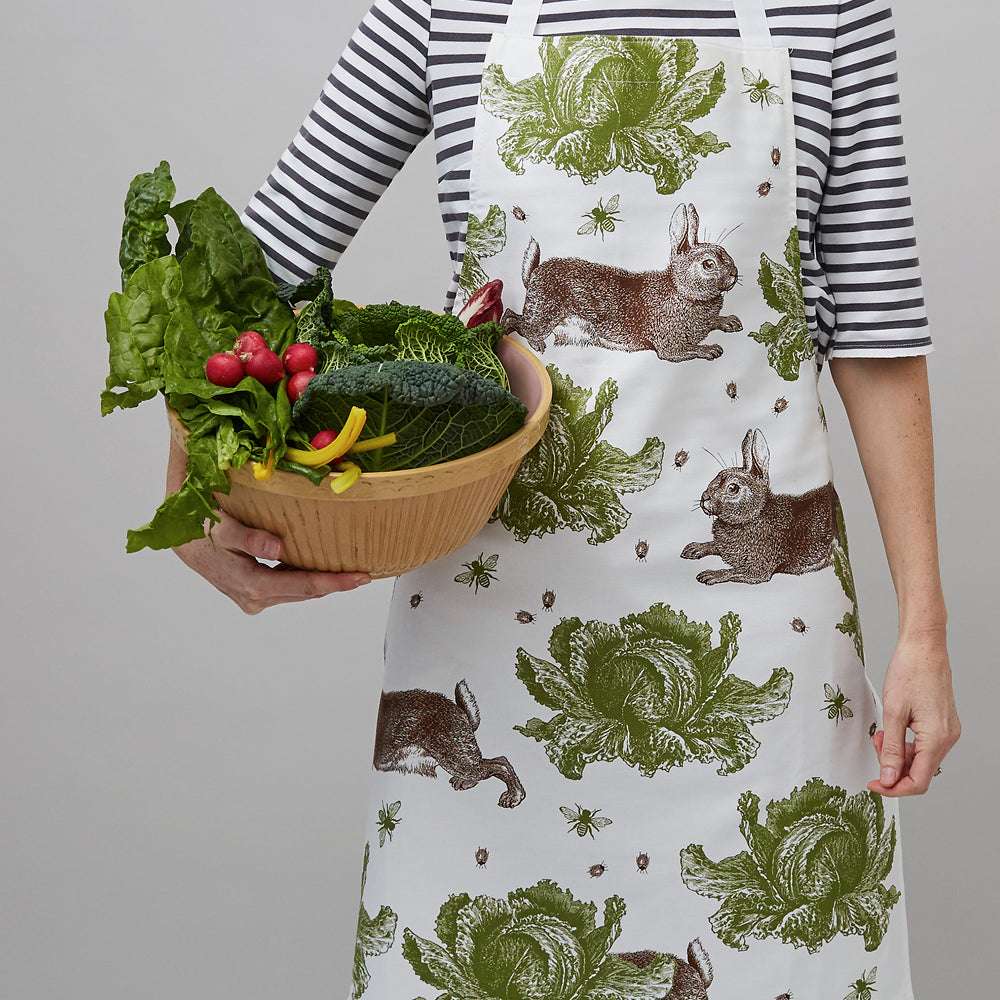 Eine Person trägt eine weiße Schürze mit grünen Kohl- und braunen Kaninchenmotiven und hält eine Schale mit frischem Gemüse.