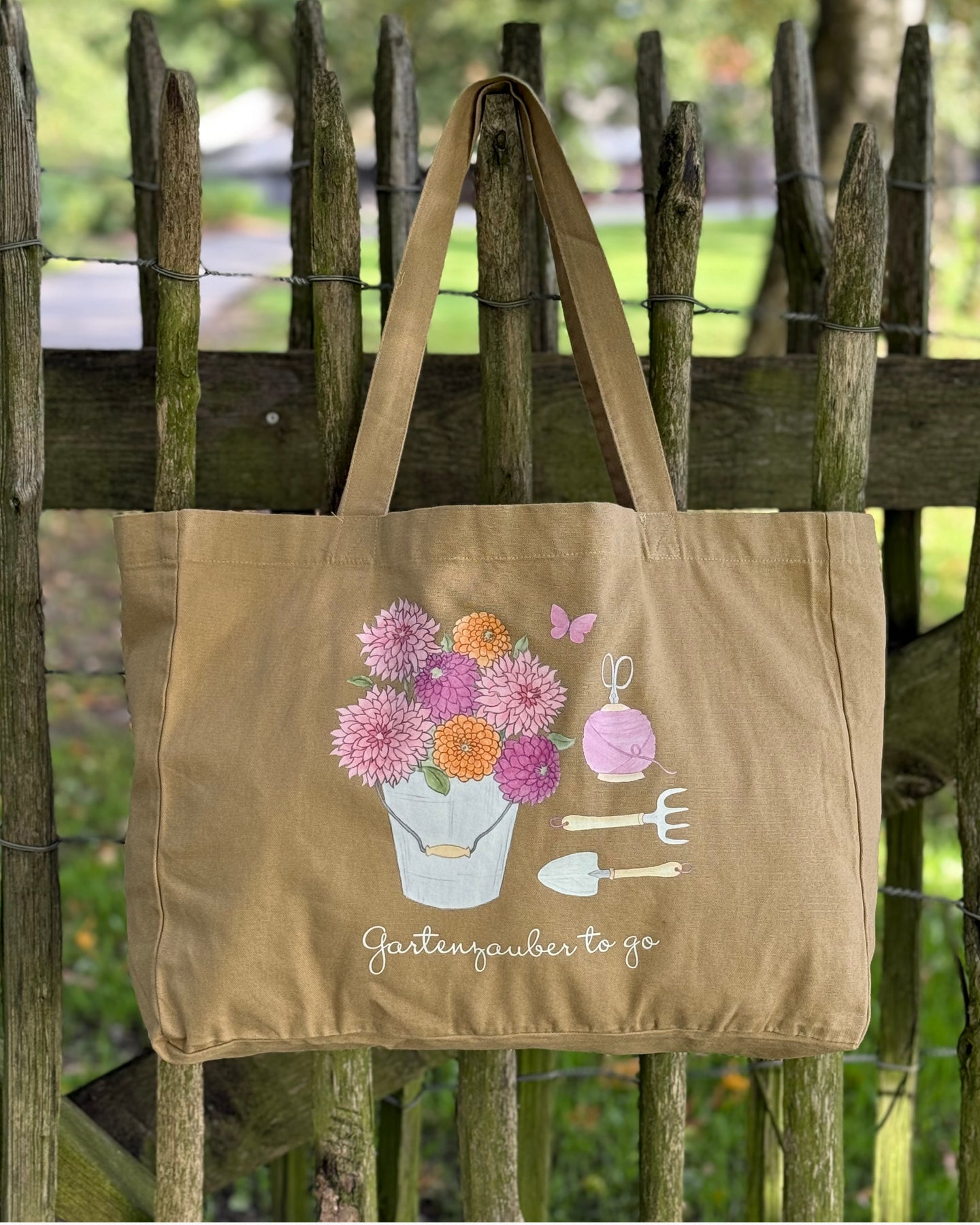 „Baumwoll-Shopper ‚Gartenzauber to go‘ im Washed-Look, mit handgezeichneten Gartenzauber-Motiven, hängend am Holzgeländer im Grünen.“