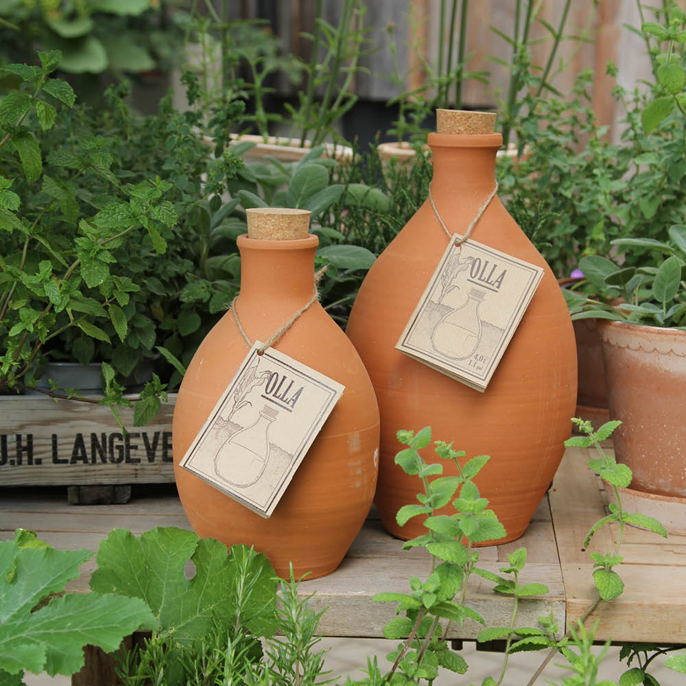 Zwei Terrakotta-Tonkrüge mit Korkverschlüssen, umgeben von grünen Pflanzen in einem Garten. Jeder Krug hat ein Etikett.