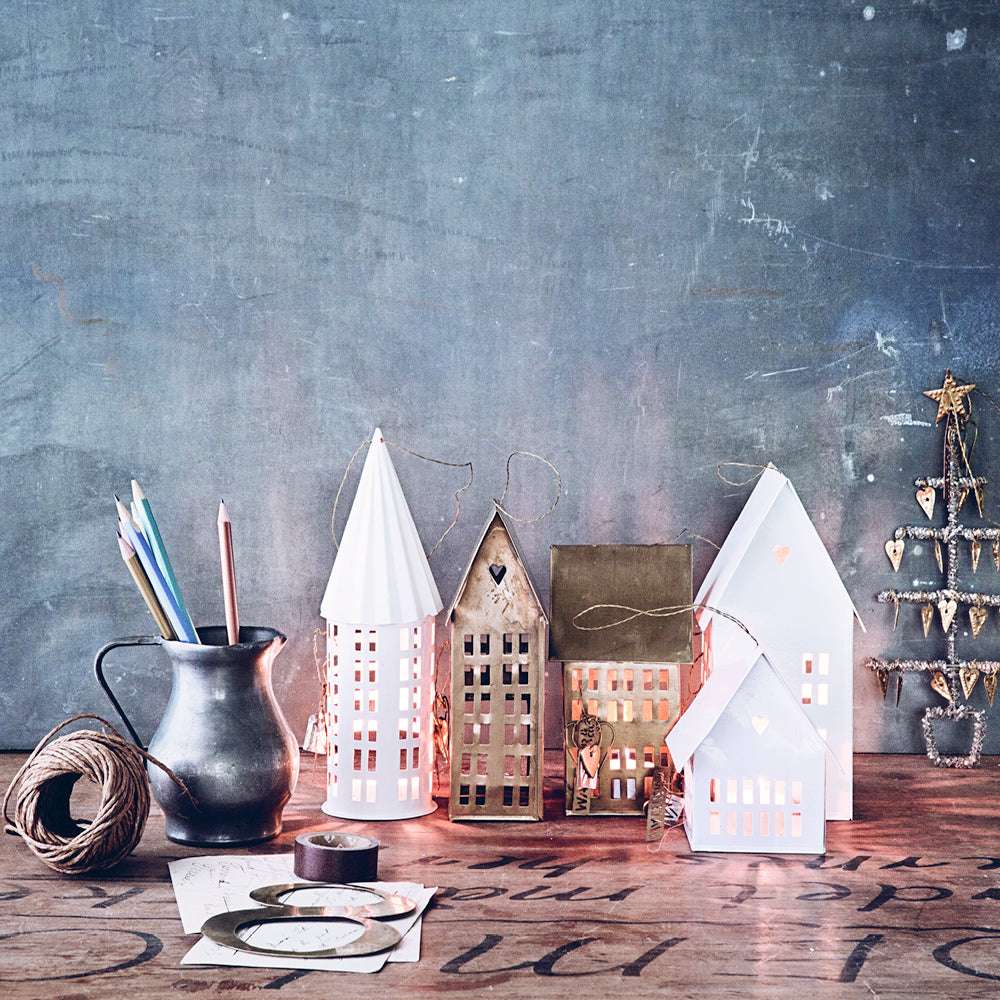 Miniaturhaus aus Messing mit Lichteffekt auf Holztisch, umgeben von dekorativen Accessoires und kreativem Hintergrund.